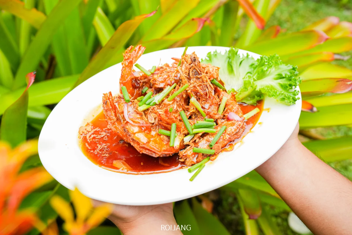 จานกุ้งปรุงสุกในซอสสีแดง ตกแต่งด้วยต้นหอมและผักกาดหอม ถูกยกมาเสิร์ฟกลางแจ้งท่ามกลางต้นไม้สีเขียว—เป็นคำที่สดชื่นสมบูรณ์แบบจากโอโซนคาเฟ่.