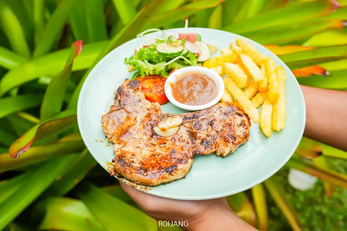 จานหนึ่งที่ร้านโอโซนคาเฟ่ประกอบด้วยสเต็กหมูย่าง, เฟรนช์ฟราย, สลัด, มะเขือเทศย่าง, และชามเล็ก ๆ ของซอสสีน้ำตาล เสิร์ฟกลางแจ้งพร้อมต้นไม้สีเขียวเป็นฉากหลัง.