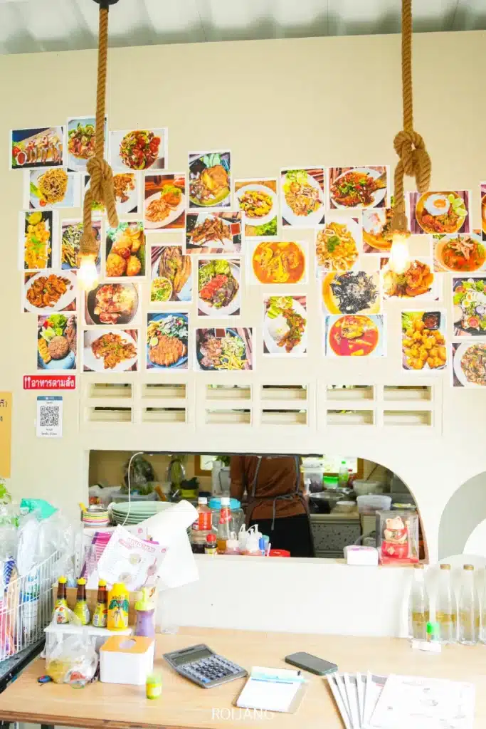 เคาน์เตอร์ร้านอาหารที่มีขวดเครื่องปรุงและเมนูอาหาร พร้อมผนังด้านหลังตกแต่งด้วยภาพถ่ายอาหารหลากหลายชนิดภายใต้หลอดไฟแขวน สร้างบรรยากาศอบอุ่นที่ Ozone Cafe