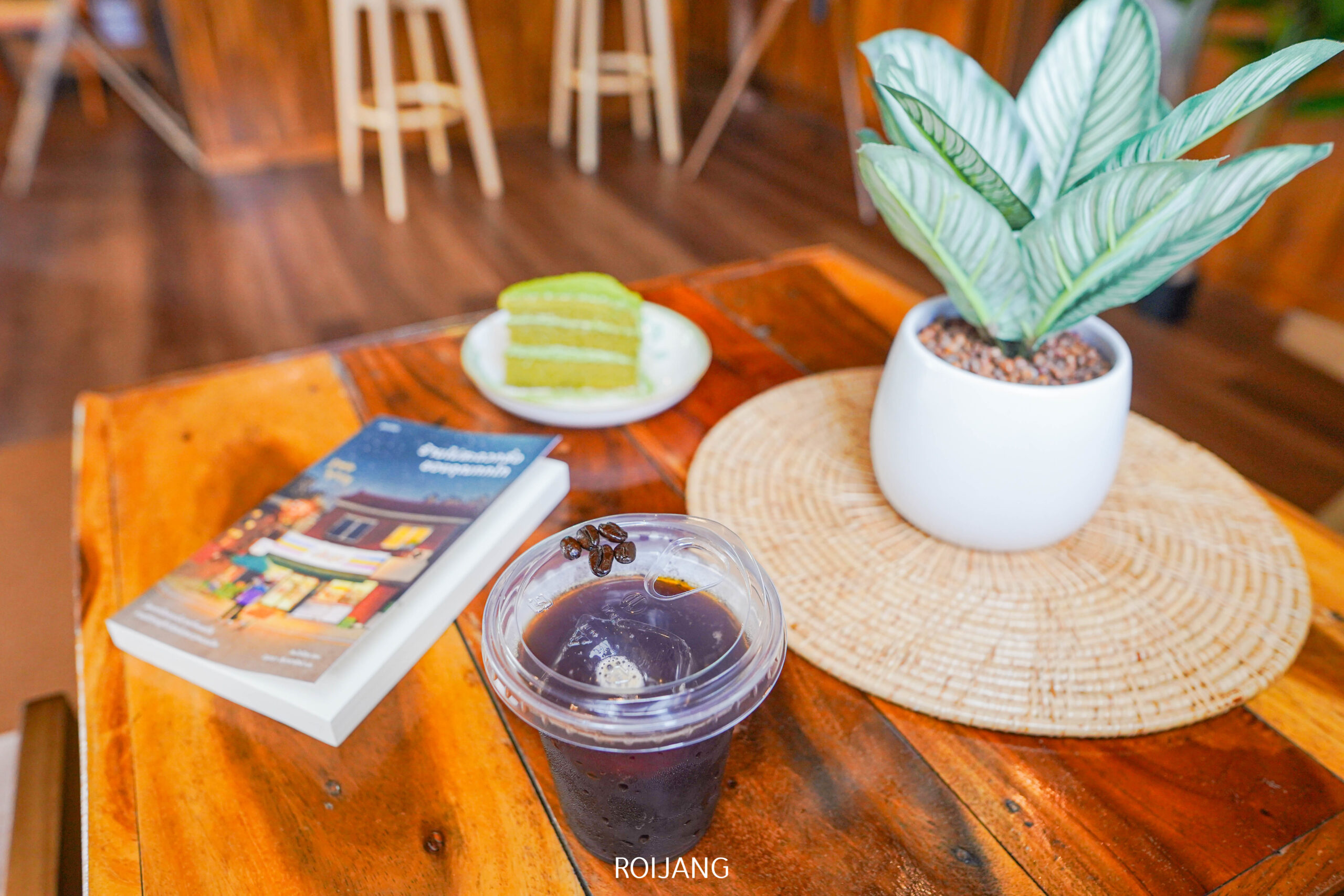 แก้วพลาสติกใส่น้ำแข็งกาแฟ, หนังสือ, จานที่มีเค้กสีเขียวสองชิ้น, และต้นไม้ในกระถางบนโต๊ะไม้ สร้างบรรยากาศอบอุ่นภายในที่ ศิ-วรา คาเฟ่.