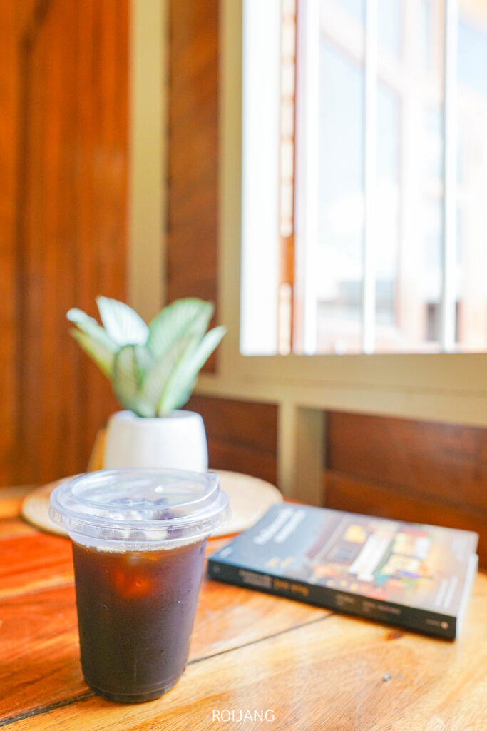 แก้วพลาสติกใส่น้ำแข็งกาแฟจาก ศิ-วรา คาเฟ่ ตั้งอยู่บนโต๊ะไม้ข้าง ๆ หนังสือและต้นไม้กระถางเล็ก ๆ โดยมีแสงแดดส่องผ่านหน้าต่างเป็นฉากหลัง.