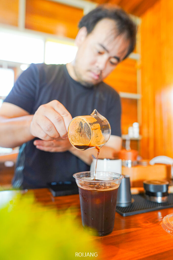 ชายคนหนึ่งกำลังรินกาแฟจากเหยือกแก้วลงในแก้วพลาสติกที่เต็มไปด้วยน้ำแข็งบนเคาน์เตอร์ไม้ที่ ศิ-วรา คาเฟ่.