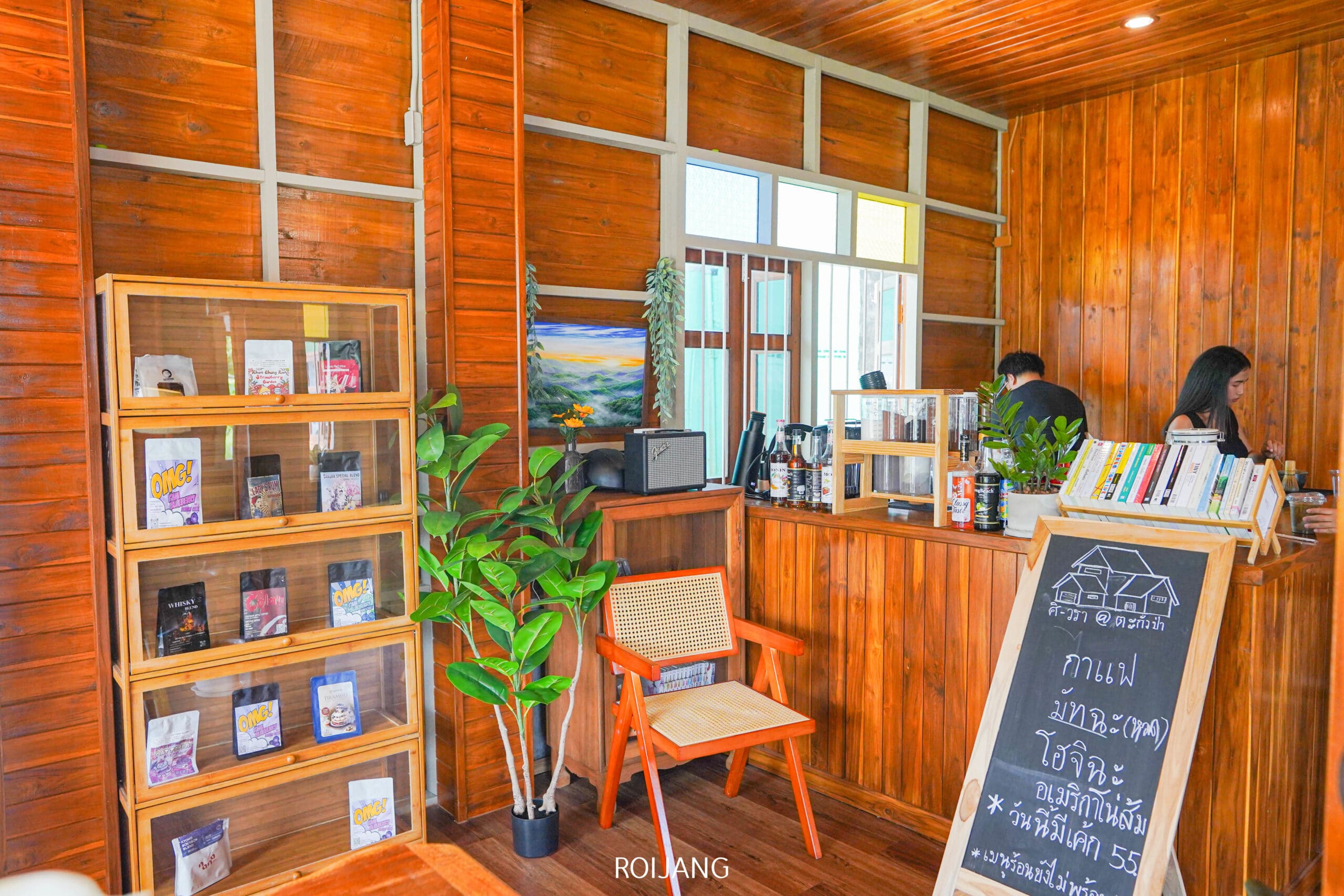บรรยากาศภายในคาเฟ่ศิ-วราที่อบอุ่นด้วยไม้ มีชั้นวางหนังสือ ต้นไม้ เก้าอี้ เคาน์เตอร์ที่มีพนักงานสองคน และเมนูเขียนด้วยชอล์กเป็นภาษาไทย.