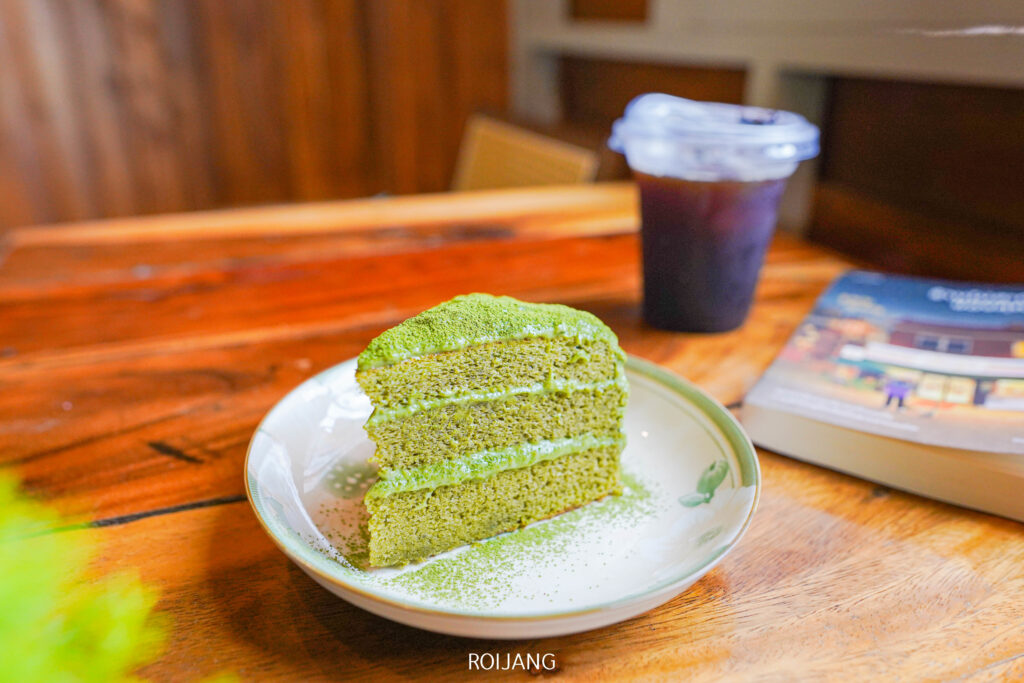 เค้กชั้นชาเขียวชิ้นหนึ่งวางอยู่บนจานบนโต๊ะไม้ที่ ศิ-วรา คาเฟ่ ข้างแก้วพลาสติกใส่น้ำแข็งกาแฟและหนังสือที่ปิดอยู่.