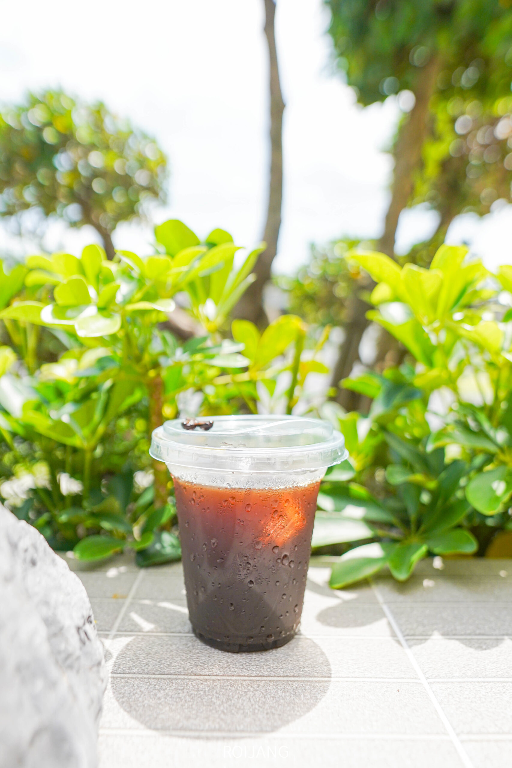 แก้วพลาสติกใส่น้ำแข็งกาแฟดำจาก ศิ-วรา คาเฟ่ พร้อมฝาครอบ ตั้งอยู่บนพื้นกระเบื้องกลางแจ้ง ล้อมรอบด้วยต้นไม้สีเขียวและแสงแดดสดใส.