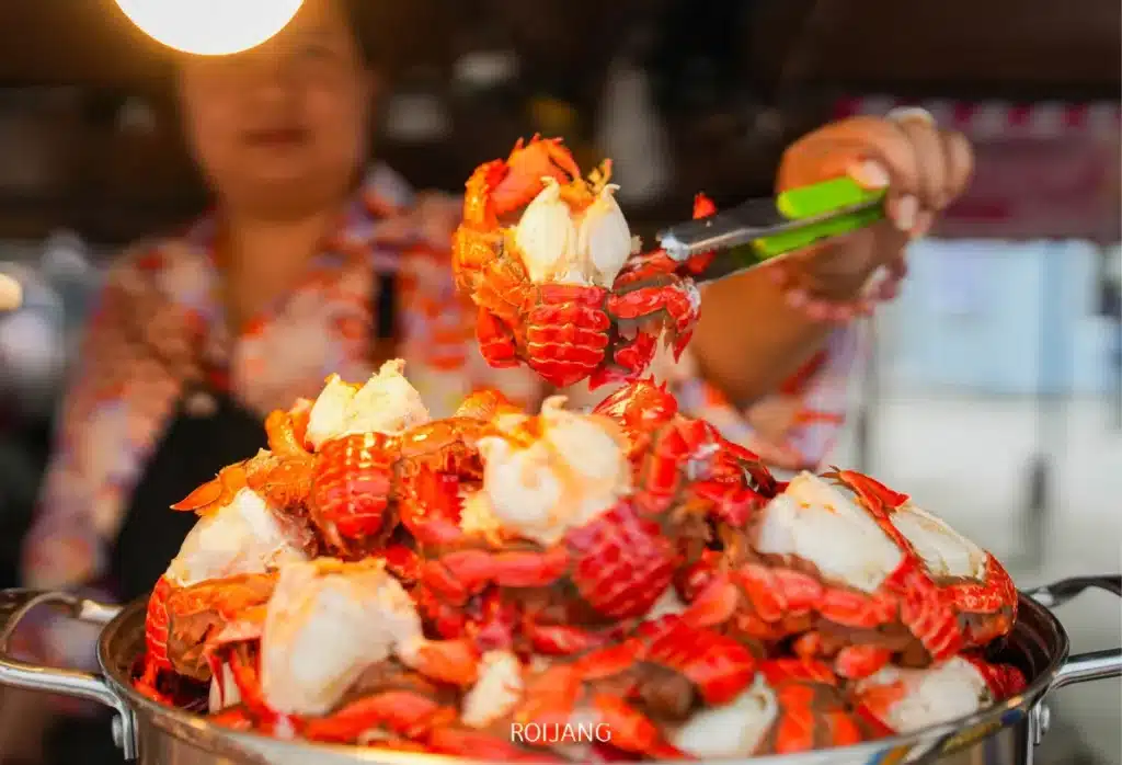 ที่ หลาดเพลินเป๋า ผู้คนใช้คีมคีบกุ้งมังกรสีแดงจากกองกุ้งมังกรขนาดใหญ่ที่วางเรียงกันในภาชนะโลหะ.