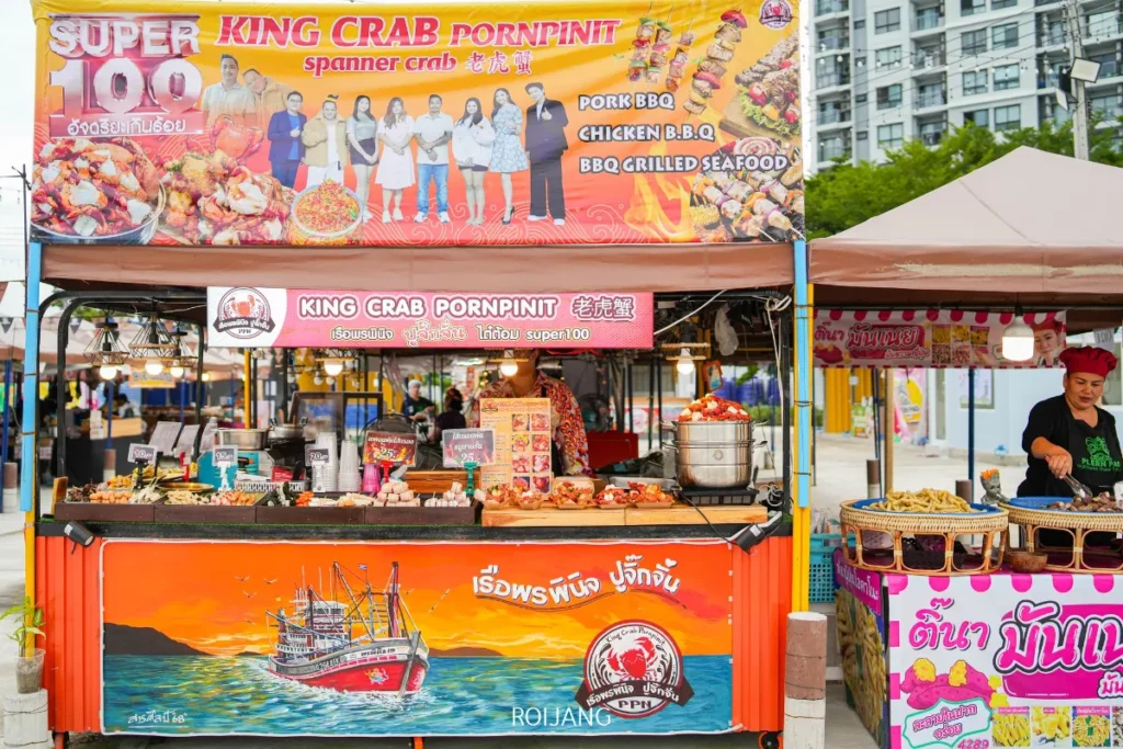 ร้านขายอาหารริมทางชื่อ "คิงแครบ พิมพ์ปิ่น" ที่ตลาดเพลินเป๋า จำหน่ายหมูบาร์บีคิว ไก่ อาหารทะเล และปู โดยมีป้ายสีสันสดใสและพ่อค้าที่เป็นมิตรอยู่หลังเคาน์เตอร์.