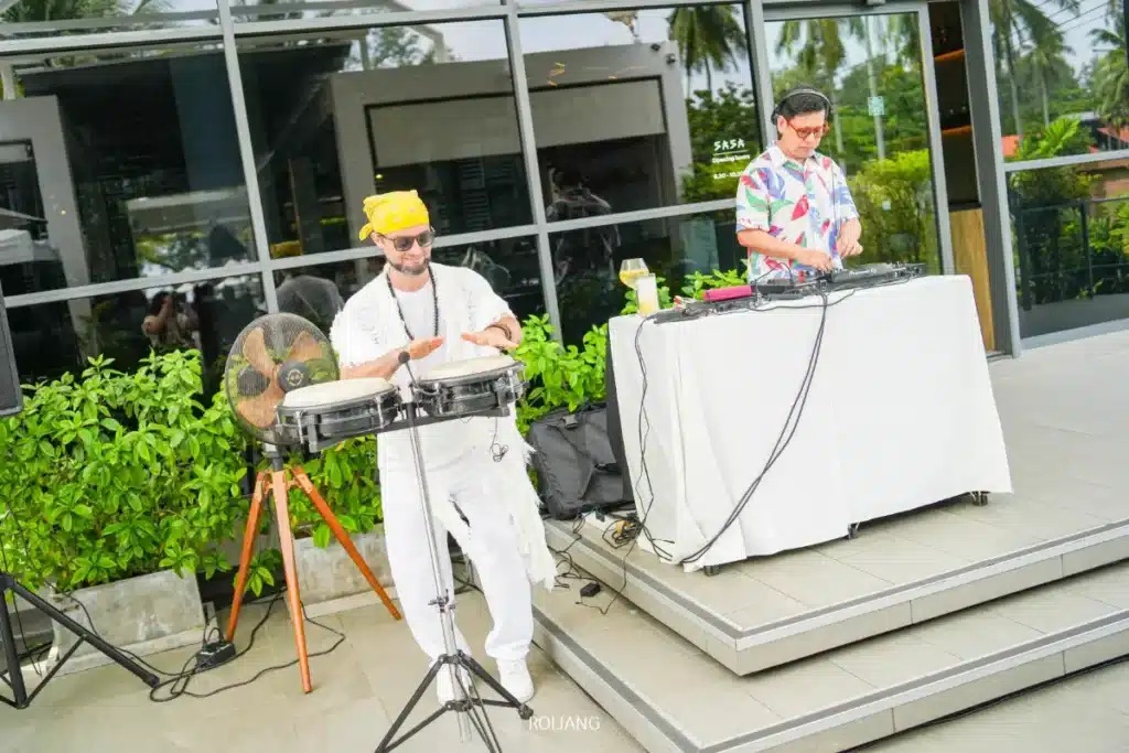 นักดนตรีสองคนแสดงดนตรีกลางแจ้งที่โรงแรม Meliá Phuket; คนหนึ่งเล่นกลองบองโกในขณะที่อีกคนหนึ่งควบคุมอุปกรณ์ดีเจบนเวทีใกล้กับอาคารที่มีหน้าต่างบานใหญ่ นำพลังชีวิตที่สดใสมาสู่ Soul of Saturday Brunch.