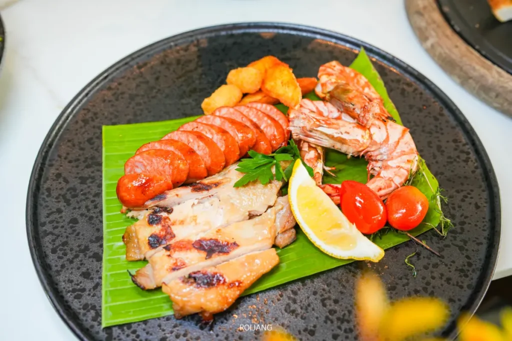 จานสีดำที่มีไก่ย่าง, ไส้กรอกหั่น, กุ้ง, มะเขือเทศเชอร์รี่, มะนาวฝาน, และของทอดบนใบตอง ถ่ายทอดรสชาติที่สดใสของ "Soul of Saturday Brunch" ที่โรงแรม Meliá Phuket.