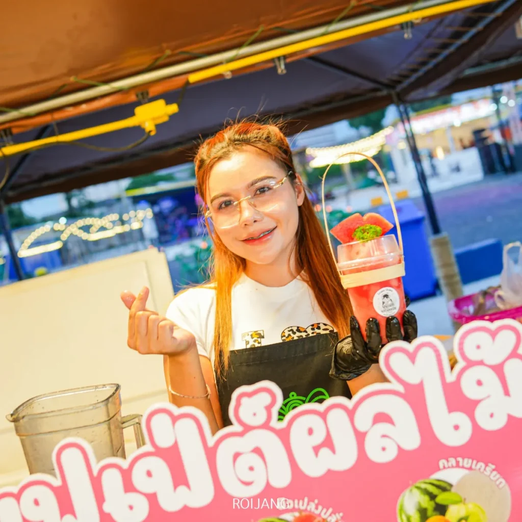 ผู้หญิงสวมถุงมือและแว่นตา ยืนอยู่หลังแผงขายของที่ หลาดเพลินเป๋า ถือของหวานที่มีสตรอว์เบอร์รี่อยู่ด้านหน้า ป้ายสีสันสดใสที่มีข้อความภาษาไทย.