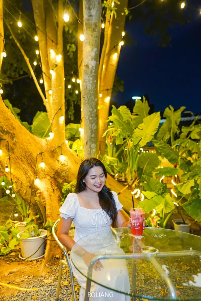 หญิงสาวในชุดสีขาวนั่งอยู่ที่โต๊ะกระจกกลางแจ้งในยามค่ำคืน ล้อมรอบด้วยไฟประดับและต้นไม้สีเขียว มีเครื่องดื่มสีแดงอยู่ตรงหน้าเธอ กำลังเพลิดเพลินกับบรรยากาศเงียบสงบและแสงสลัวของยามเย็น.