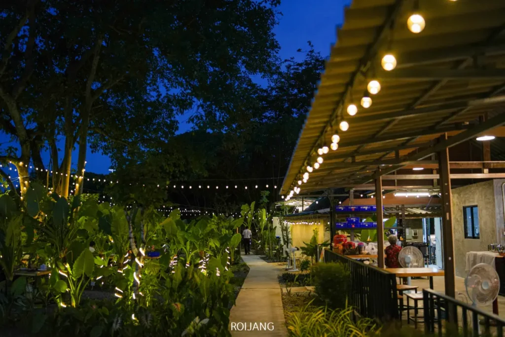ร้านอาหารกลางแจ้งยามพลบค่ำพร้อมไฟประดับ ต้นไม้สีเขียว และทางเดินที่มีหลังคาคลุม มีคนยืนอยู่ใกล้ทางเดินใต้ต้นไม้ที่ส่องแสงสว่างและท้องฟ้ายามเย็นสีฟ้า เพลิดเพลินกับบรรยากาศอบอุ่นของร้าน.