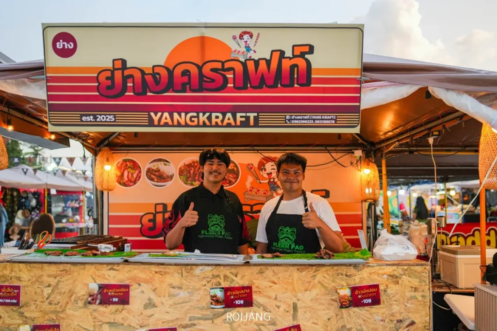 ชายสองคนยืนยิ้มและชูนิ้วโป้งอยู่หลังเคาน์เตอร์ของร้านอาหารที่มีป้ายชื่อ "YANGKRAFT" ที่ตลาดนัดเพลินเป๋า ซึ่งเป็นตลาดกลางแจ้ง บนเคาน์เตอร์มีอาหารหลากหลายชนิดและราคาแสดงอยู่.
