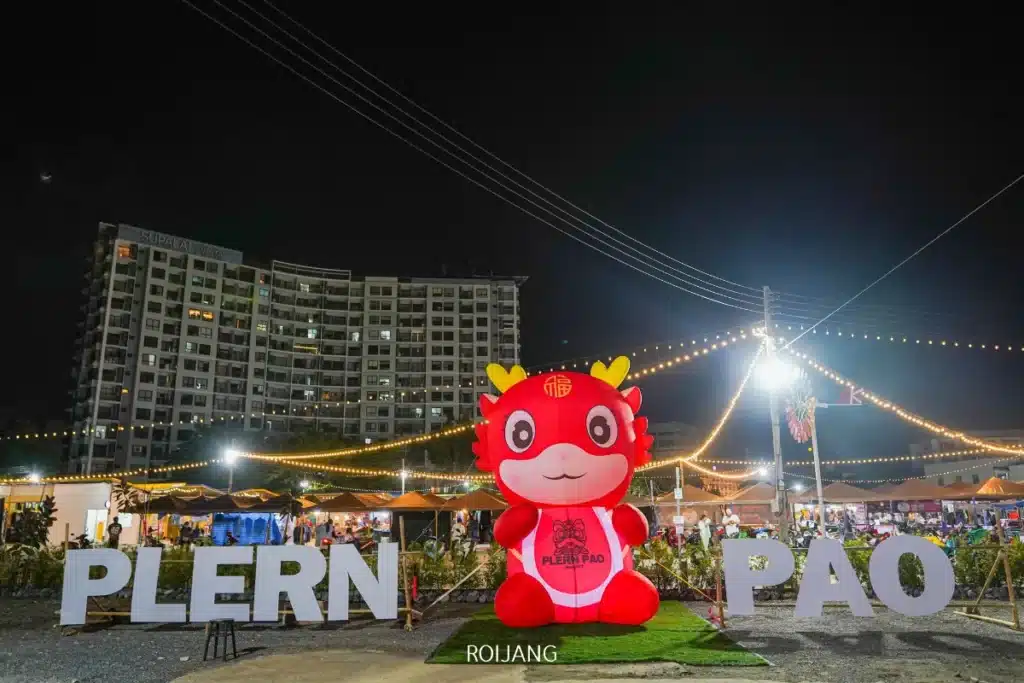 มาสคอตการ์ตูนสีแดงขนาดใหญ่ยืนอยู่ระหว่างตัวอักษร "PLERN PAO" ที่ตลาดนัดกลางคืน "หลาดเพลินเป๋า" โดยมีไฟส่องสว่าง เต็นท์สีสันสดใส และอาคารอพาร์ตเมนต์สูงตระหง่านเป็นฉากหลัง.