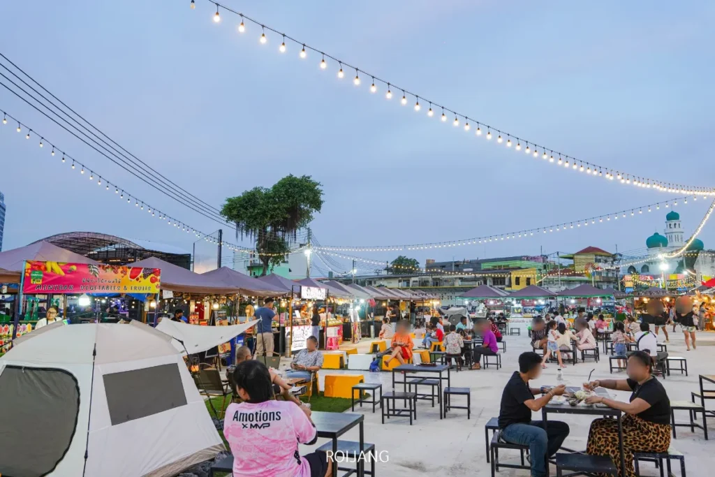 ตลาดนัดกลางคืนกลางแจ้ง หลาดเพลินเป๋า พร้อมไฟประดับ, ร้านอาหาร, และผู้คนนั่งที่โต๊ะกินอาหารและสังสรรค์. เต็นท์และป้ายสีสันสดใสสามารถมองเห็นได้ในพื้นหลัง.