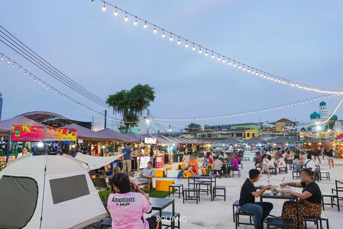 ตลาดนัดกลางคืนกลางแจ้ง หลาดเพลินเป๋า พร้อมไฟประดับ, ร้านอาหาร, และผู้คนนั่งที่โต๊ะกินอาหารและสังสรรค์. เต็นท์และป้ายสีสันสดใสสามารถมองเห็นได้ในพื้นหลัง.