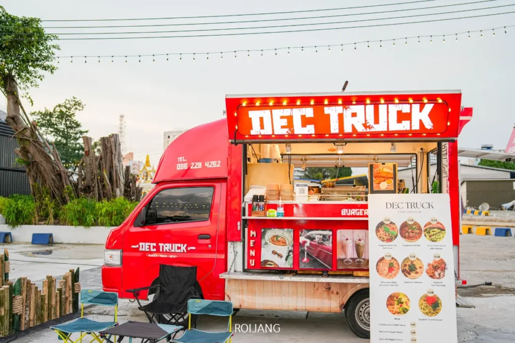 รถขายอาหารสีแดงที่มีป้ายเขียนว่า "DEC TRUCK" จอดอยู่กลางแจ้งที่ หลาดเพลินเป๋า โดยมีป้ายเมนูและพื้นที่นั่งรับประทานอาหารตั้งอยู่ด้านหน้าบนพื้นคอนกรีต.