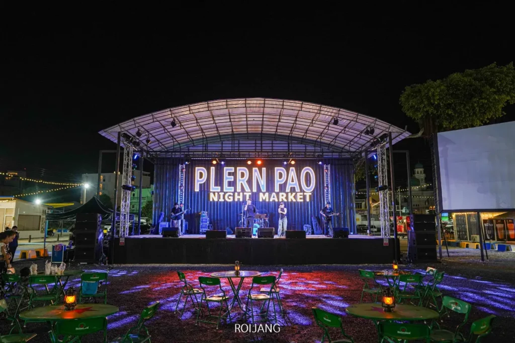 เวทีกลางแจ้งที่ตลาดนัดเพลินเป๋าในเวลากลางคืน มีนักดนตรีแสดงภายใต้แสงไฟสว่างจ้า; มีเก้าอี้สีเขียวและโต๊ะว่างถูกจัดวางไว้หน้าเวทีในเวลากลางคืน.