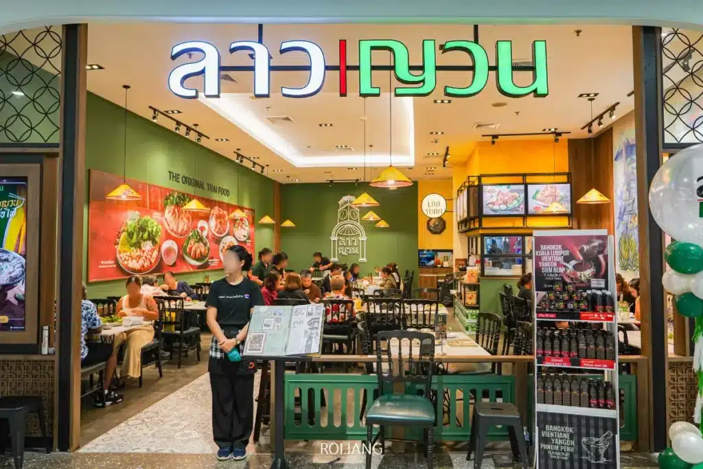ภาพด้านหน้าของร้านอาหารไทยที่สว่างไสว ชื่อว่า ลาวญวน มีลูกค้าทานอาหารอยู่ภายใน พนักงานถือเมนูอยู่ที่ทางเข้า และมีป้ายสีสันสดใสอยู่เหนือประตู.