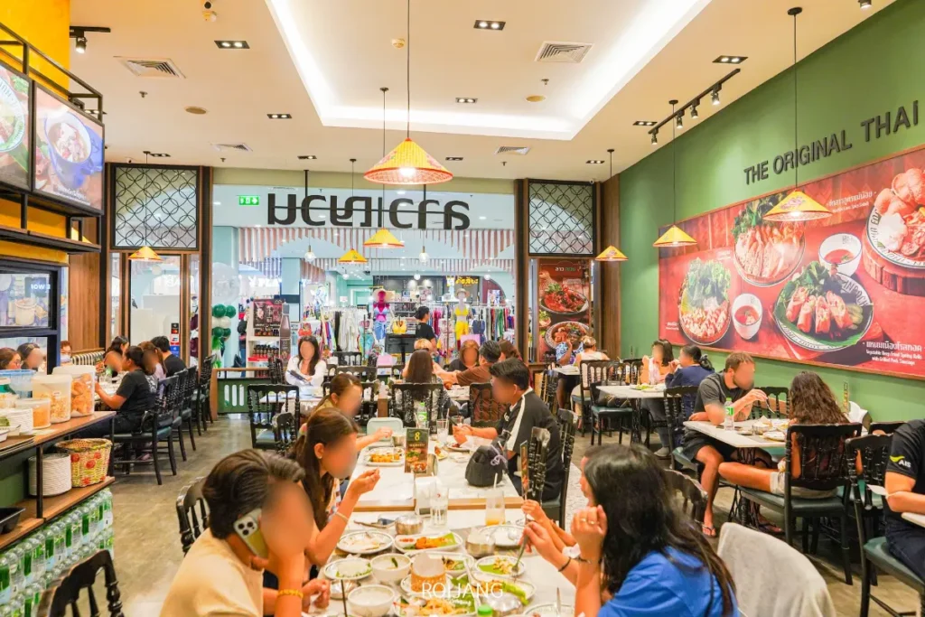 ร้านอาหารลาวญวนไทยที่คึกคัก มีลูกค้าเพลิดเพลินกับอาหารที่โต๊ะ จานอาหารสีสันสดใสจัดแสดงบนผนัง และมองเห็นทางเดินในห้างสรรพสินค้าผ่านทางเข้าของร้านอาหาร.