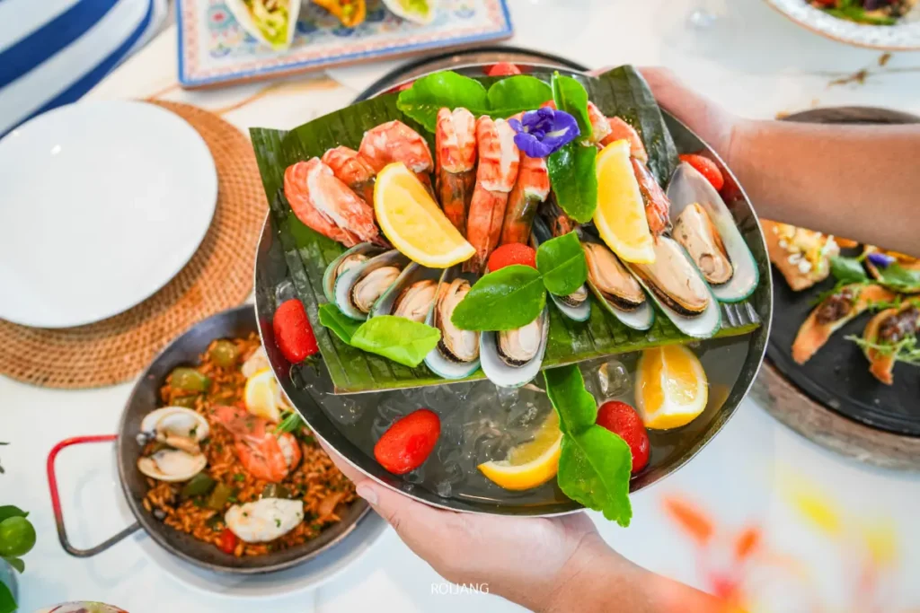 จานอาหารทะเลสดใหม่ที่ Soul of Saturday Brunch ของ Meliá Phuket ประกอบด้วยกุ้ง, หอยแมลงภู่, และมะนาวฝานเสิร์ฟบนน้ำแข็ง ตกแต่งด้วยใบเขียวและดอกไม้ที่รับประทานได้.