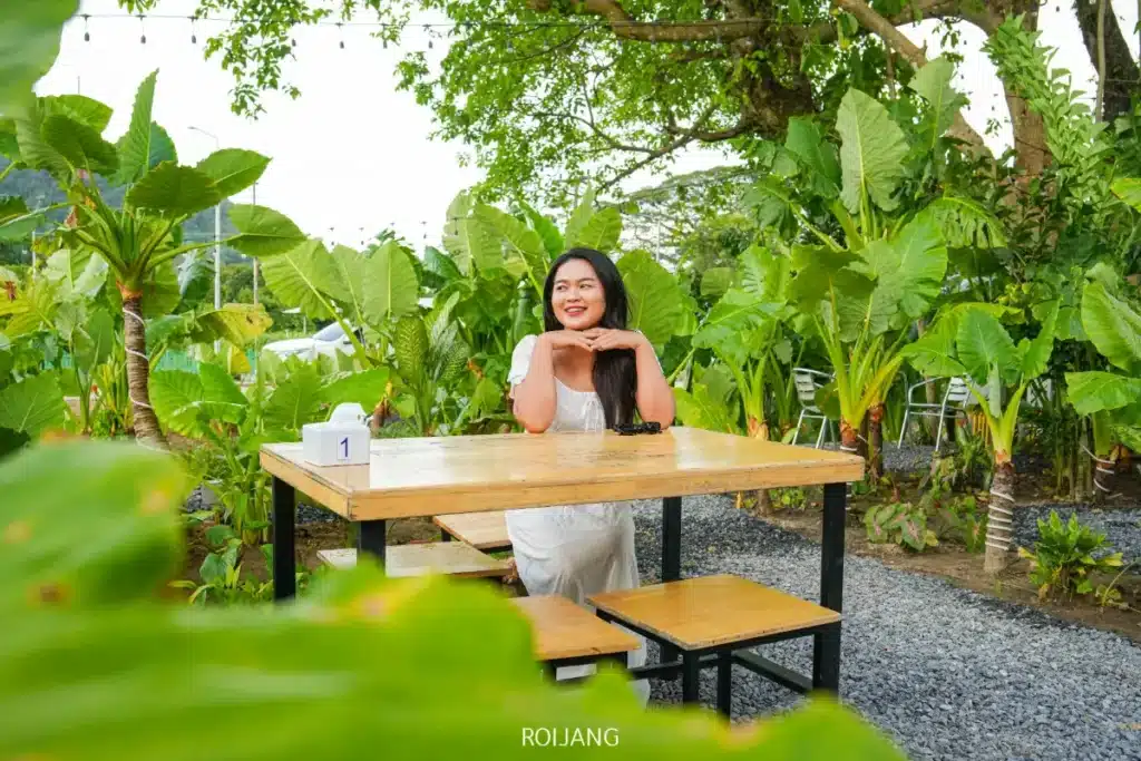 ผู้หญิงในชุดสีขาว นั่งอยู่คนเดียวที่โต๊ะไม้กลางแจ้ง ล้อมรอบด้วยต้นไม้สีเขียวชอุ่ม ยิ้มอยู่ใต้คางของเธอ มือทั้งสองข้างอยู่ใต้คางของเธอ แสดงถึงความเงียบสงบและเสน่ห์ที่แอบแฝง.