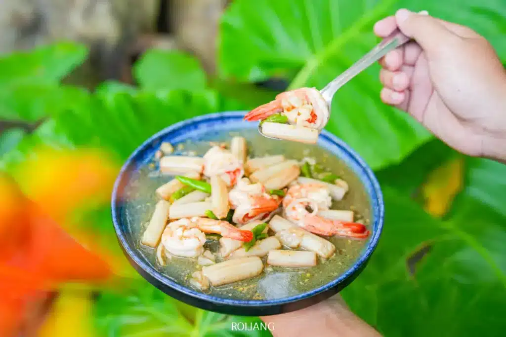 ชามสีน้ำเงินเต็มไปด้วยกุ้ง หน่อไม้ และผักในน้ำซุปถูกถือไว้ในมือข้างหนึ่ง ในขณะที่มืออีกข้างหนึ่งยกช้อนที่ตัก แอบแอ ขึ้นมา ใบไม้สีเขียวอยู่เบื้องหลัง.