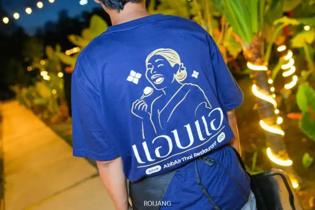 บุคคลสวมเสื้อเชิ้ตสีน้ำเงินที่มีภาพกราฟิกของผู้หญิงกำลังกินและมีข้อความภาษาไทยว่า "แอบแอ" ยืนอยู่กลางแจ้งใกล้กับต้นไม้ที่มีแสงไฟในเวลากลางคืน.