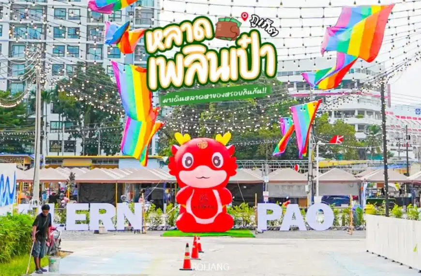 รูปปั้นมาสคอตการ์ตูนสีแดงตั้งอยู่หน้าตัวอักษรสีขาวขนาดใหญ่และธงสีรุ้งที่งานตลาดกลางแจ้ง หลาดเพลินเป๋า ในบรรยากาศเมือง มีข้อความและของตกแต่งภาษาไทยอยู่ด้านบน.