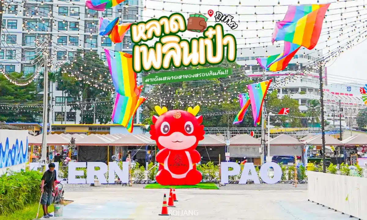 รูปปั้นมาสคอตการ์ตูนสีแดงตั้งอยู่หน้าตัวอักษรสีขาวขนาดใหญ่และธงสีรุ้งที่งานตลาดกลางแจ้ง หลาดเพลินเป๋า ในบรรยากาศเมือง มีข้อความและของตกแต่งภาษาไทยอยู่ด้านบน.