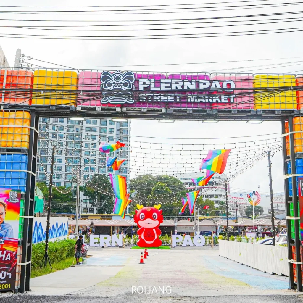 ทางเข้าตลาดเพลินเป่า หรือ หลาดเพลินเป๋า มีป้ายสีสันสดใส ธงสีรุ้ง ตุ๊กตา mascot สีแดงขนาดใหญ่ และแผงขายของในตลาดที่คึกคักเป็นฉากหลัง.