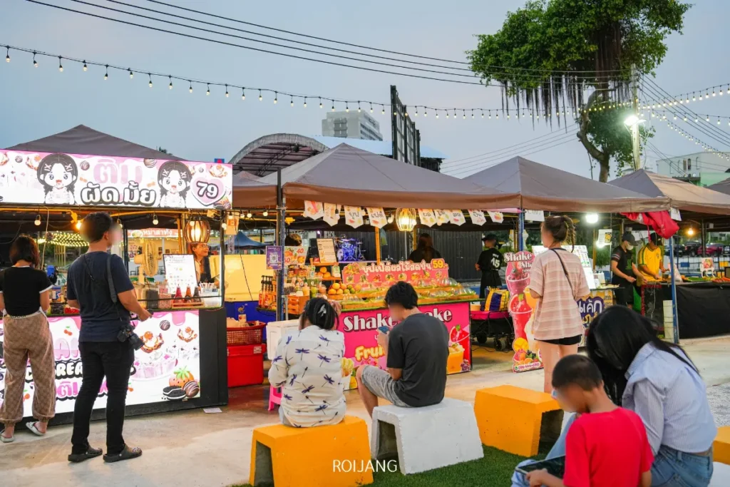 ผู้คนกำลังช้อปปิ้งและผ่อนคลายที่ หลาดเพลินเป๋า, ตลาดกลางคืนกลางแจ้งที่รายล้อมไปด้วยร้านอาหาร, ไฟประดับ, และป้ายสีสันสดใส.