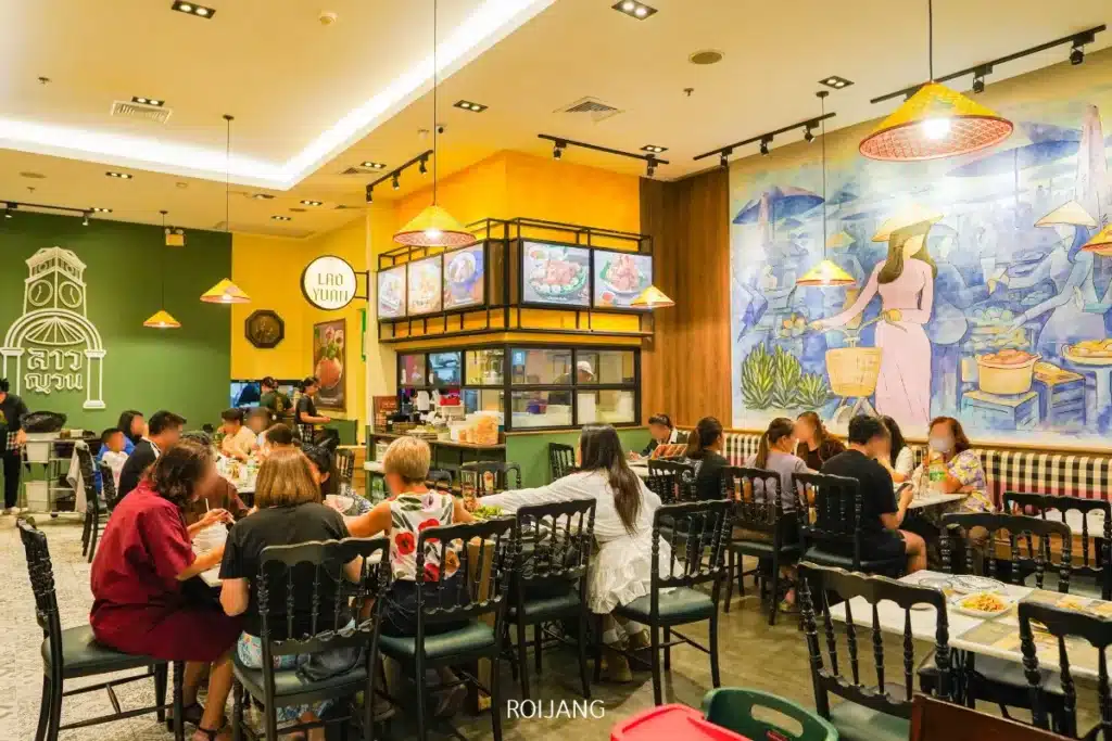 ร้านอาหารที่คึกคัก ลาวญวน มีลูกค้าที่นั่งอยู่ที่โต๊ะ ศิลปะบนผนังสีสันสดใสที่วาดภาพตลาด และไฟเพดานที่สว่างไสว.