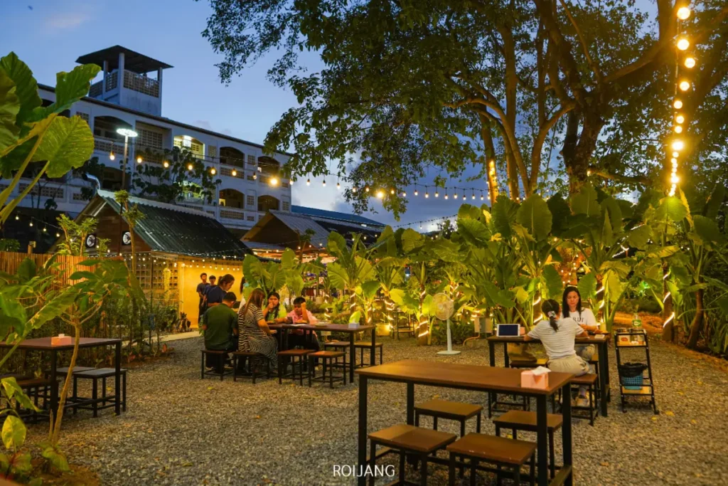 ร้านอาหารกลางแจ้งที่มีไฟประดับแบบสายไฟ, ต้นไม้เขียวชอุ่ม, และกลุ่มคนนั่งรับประทานอาหารที่โต๊ะในตอนเย็น. บรรยากาศอบอุ่นและสบาย ๆ, พร้อมตึกใหญ่ที่ส่องสว่างอยู่เบื้องหลัง.