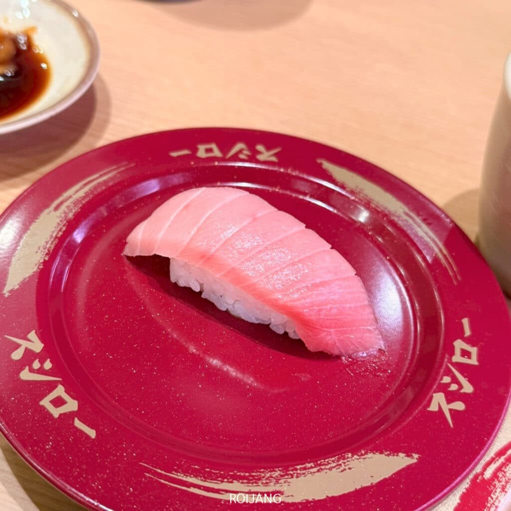 ซูชิหน้าทูน่าหนึ่งชิ้นวางบนจานสีแดงพร้อมตัวหนังสือภาษาญี่ปุ่น วางอยู่บนโต๊ะสีอ่อนที่ร้าน CentralPhuketร้านอาหาร.