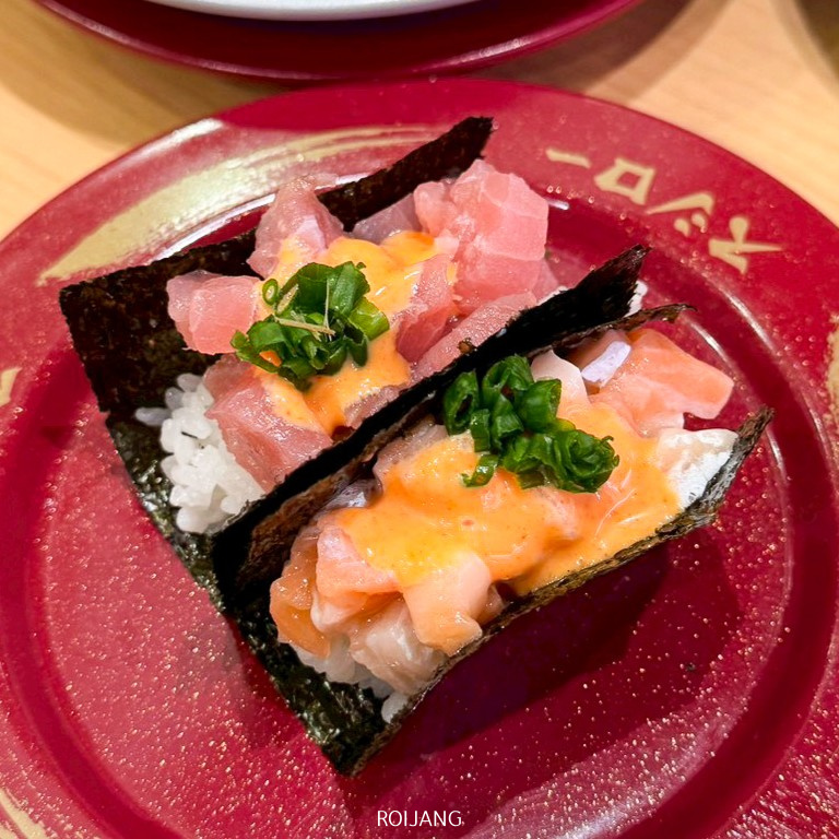 ซูชิโคนสองชิ้นที่ทำด้วยมือ โรยหน้าด้วยปลาดิบหั่นเต๋า ซอสสีส้ม และต้นหอมบนข้าว ห่อด้วยสาหร่าย เสิร์ฟบนจานสีแดงที่ร้าน CentralPhuketร้านอาหาร.