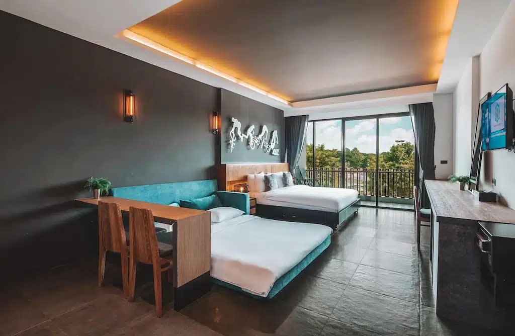 ห้องพักโรงแรมสมัยใหม่พร้อมเตียงสองเตียง โต๊ะเล็กพร้อมเก้าอี้ ทีวีติดผนัง และหน้าต่างบานใหญ่เปิดออกสู่ระเบียงที่มีวิวต้นไม้—เหมาะสำหรับการพักผ่อนหลังจากสำรวจสถานที่ท่องเที่ยวสระแก้ว.