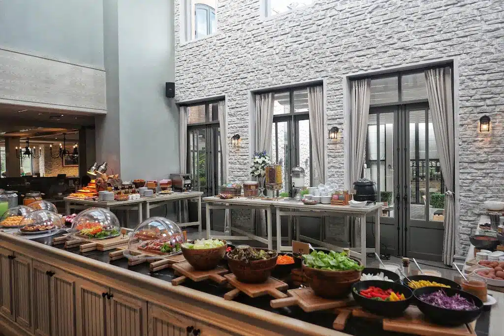 การจัดบุฟเฟ่ต์ในพื้นที่รับประทานอาหารที่สว่างและทันสมัยที่ โรงแรมเขาใหญ่ มีสลัดหลากหลาย, ท็อปปิ้ง, และอาหารที่ปิดฝาไว้เรียงรายบนเคาน์เตอร์ไม้.
