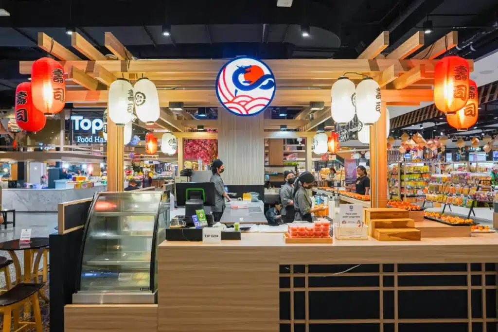 ร้านซูชิในซูเปอร์มาร์เก็ตที่ร้านอาหารในเซ็นทรัลภูเก็ต มีโคมไฟ, โลโก้ปลา, ตู้โชว์, และพนักงานกำลังเตรียมอาหารอยู่หลังเคาน์เตอร์.