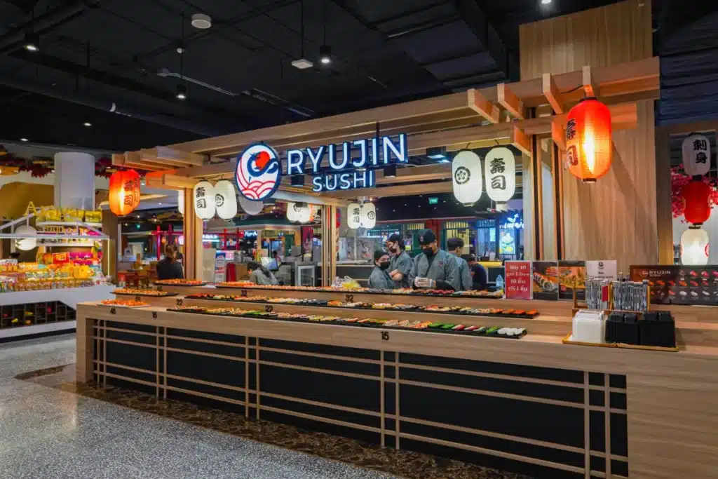 ร้านซูชิ ริวจิน ซูชิ ซึ่งเป็นเคาน์เตอร์ซูชิที่ ร้านอาหารเซ็นทรัลภูเก็ต แสดงซูชิโรลหลากหลายชนิด โดยมีเชฟเตรียมอาหารอยู่ด้านหลังเคาน์เตอร์ โคมไฟญี่ปุ่นและป้ายตกแต่งร้าน.