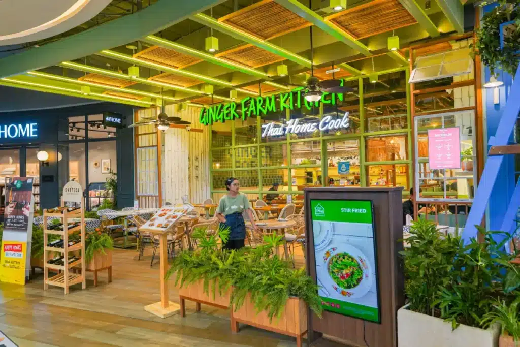 บุคคลหนึ่งยืนอยู่ด้านนอกของ Ginger Farm Kitchen ร้านอาหารเซ็นทรัลภูเก็ต ที่มีผนังกระจก ที่นั่งกลางแจ้ง ป้ายไฟนีออนสีเขียว และพืชพรรณเขียวชอุ่มประดับทางเข้า.