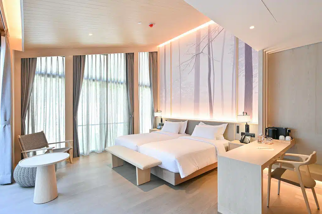 ห้องพักโรงแรมสมัยใหม่ที่ โรงแรมเขาใหญ่ พร้อมเตียงคู่สองเตียง โต๊ะทำงาน เก้าอี้มีพนักพิง โต๊ะเล็ก หน้าต่างบานใหญ่พร้อมผ้าม่าน และศิลปะบนผนังที่มีภาพเงาของต้นไม้ เหมาะสำหรับผู้ที่มองหาที่พักในสไตล์และสะดวกสบายที่ เขาใหญ่.