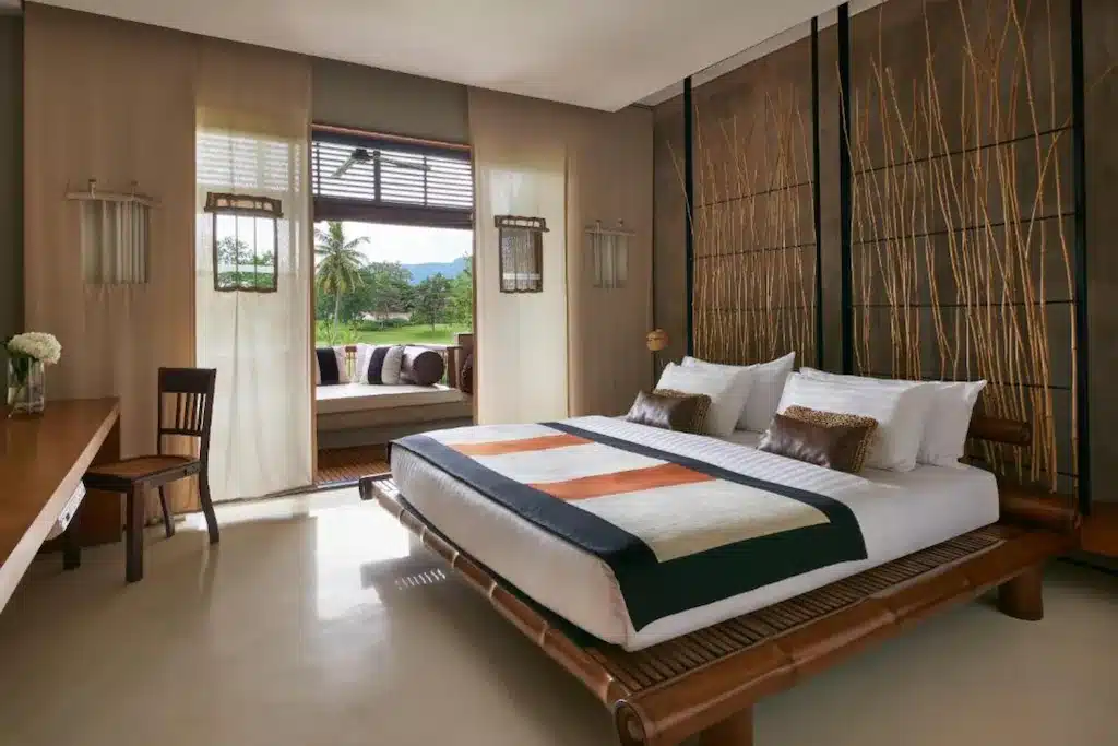 ห้องนอนสมัยใหม่ที่ โรงแรมเขาใหญ่ พร้อมเตียงขนาดใหญ่ที่มีกรอบไม้ไผ่ ตกแต่งด้วยโทนสีกลาง ๆ ผนังตกแต่งด้วยต้นหญ้าสูง โต๊ะทำงานและเก้าอี้ พร้อมที่นั่งริมหน้าต่างที่สามารถมองเห็นต้นไม้เขียวขจี.