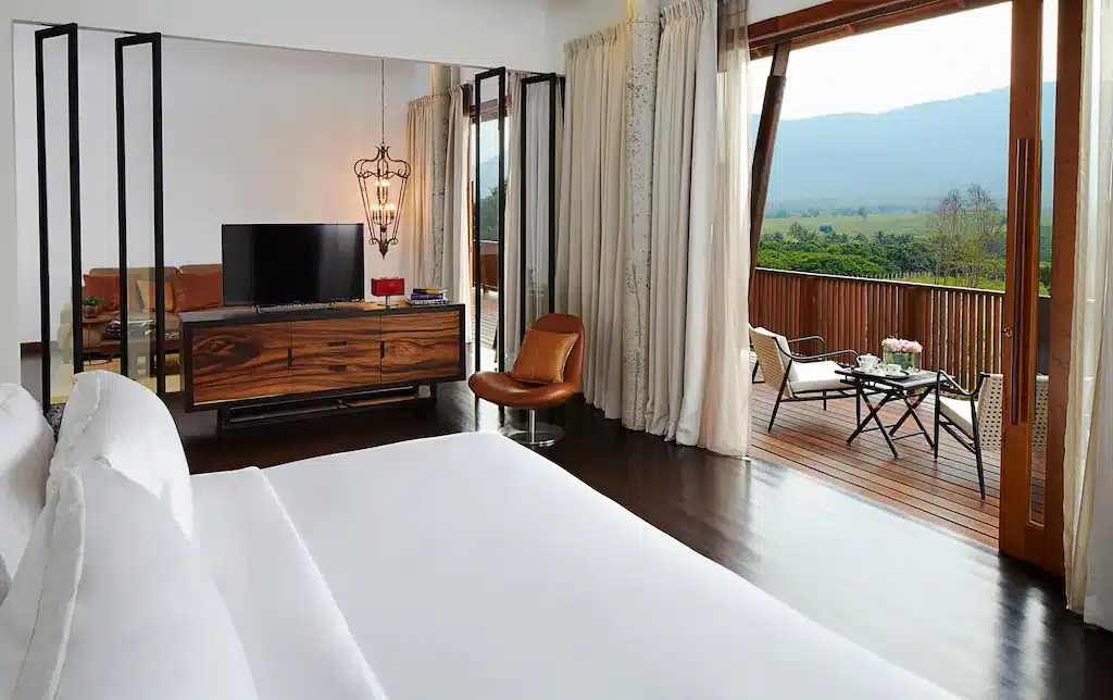 ห้องพักโรงแรมสมัยใหม่ที่ โรงแรมเขาใหญ่ พร้อมเตียงขนาดใหญ่, ทีวี, พื้นที่นั่งเล่น, และประตูบานเลื่อนเปิดออกสู่ระเบียงพร้อมเฟอร์นิเจอร์กลางแจ้งและวิวภูเขาเขียวขจี.