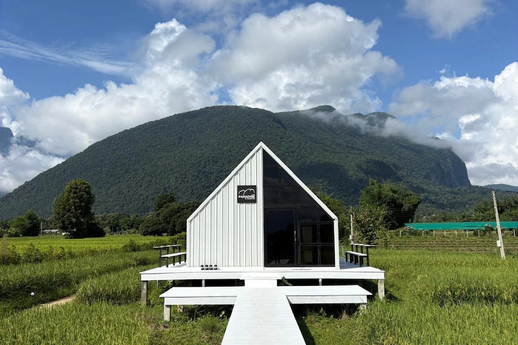 กระท่อมไม้สีขาวทรงเอ-เฟรมขนาดเล็กที่มีด้านหน้าเป็นกระจกตั้งอยู่ในทุ่งหญ้าสีเขียว ล้อมรอบด้วยภูเขาที่ปกคลุมไปด้วยป่าและท้องฟ้าที่มีเมฆครึ้ม—เหมาะสำหรับผู้ที่ต้องการ  ที่เที่ยวเชียงดาว เที่ยวเชียงดาว หรือค้นหา สถานที่ท่องเที่ยวใหม่ ๆ ในเชียงดาว.