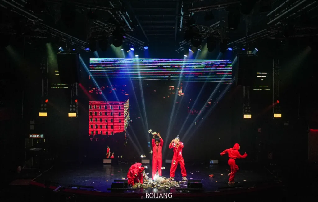 นักแสดงสี่คนในชุดจั๊มสูทสีแดงควบคุมเวทีที่ FLAME Phuket Town โดยมีแสงไฟที่ดราม่าและภาพเมืองดิจิทัลเป็นกรอบ พร้อมเศษซากที่กระจัดกระจายเพิ่มความเข้มข้นให้กับบรรยากาศ.