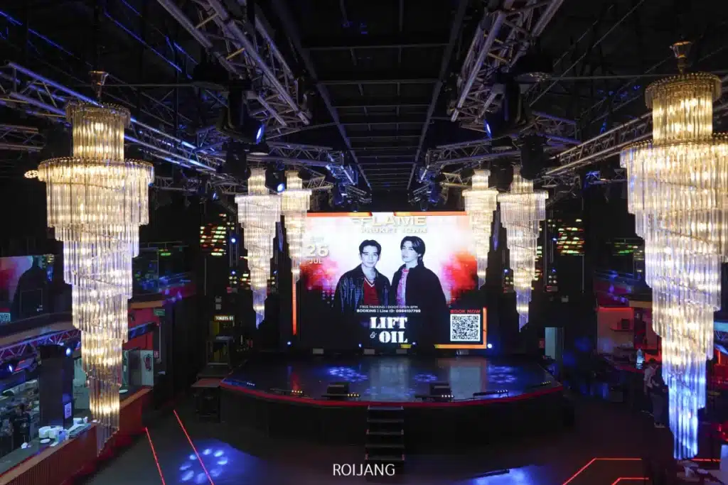 เวทีไนต์คลับที่ FLAME Phuket Town มีโคมไฟระย้าขนาดใหญ่, ไฟ LED, และจอภาพดิจิทัลที่แสดงภาพคนสองคนกำลังโปรโมตกิจกรรมพร้อมข้อความ "LIFT& OIL" และ QR code.