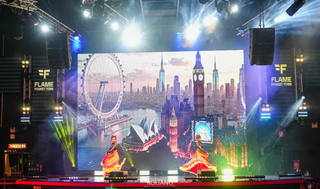 นักเต้นสองคนแสดงบนเวทีหน้าจอกดิจิตอลขนาดใหญ่ที่แสดงภาพสถานที่ท่องเที่ยวที่มีชื่อเสียงทั่วโลกที่ FLAME Phuket Town โดยมีไฟเวทีสีสันสดใสส่องสว่างให้บรรยากาศเต็มไปด้วยชีวิตชีวา.