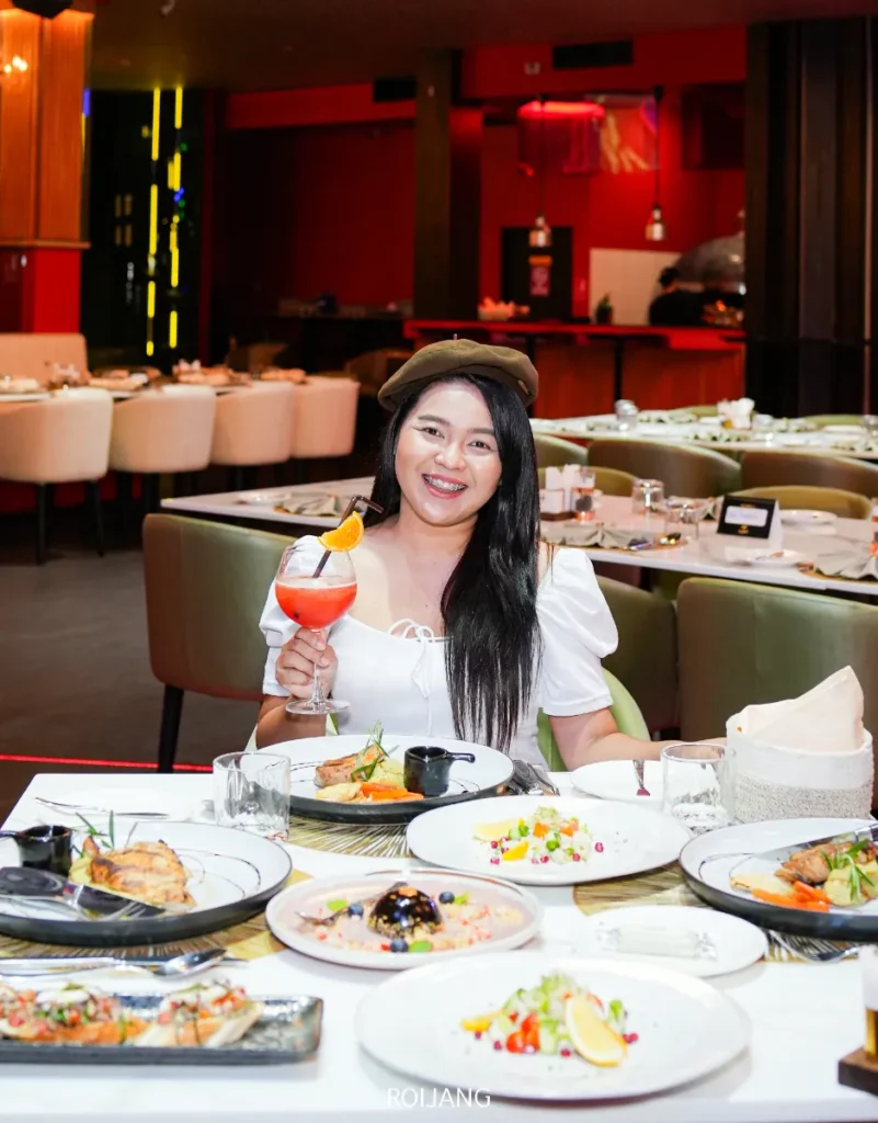 ผู้หญิงคนหนึ่งนั่งอยู่ที่โต๊ะร้านอาหาร FLAME Phuket Town พร้อมจานอาหารหลากหลายชนิด ถือเครื่องดื่มที่มีส้มประดับอยู่ ยิ้มให้กล้อง.