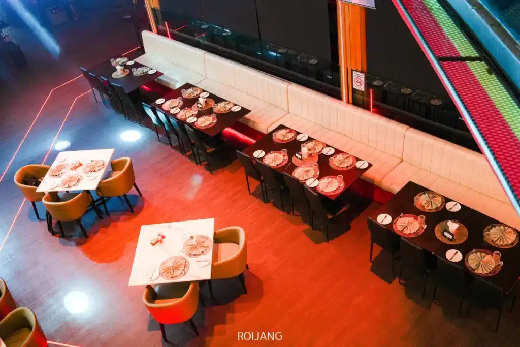 ภาพมุมสูงของร้านอาหาร FLAME Phuket Town พร้อมโต๊ะที่จัดเรียงอย่างเรียบร้อย เก้าอี้สีเบจ และม้านั่งยาวหุ้มเบาะสีขาวตามแนวกำแพงบนพื้นไม้.